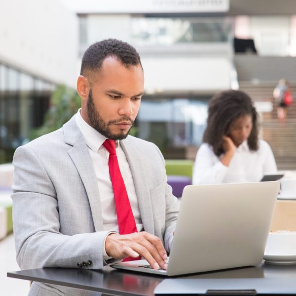 Focused business professional working on project over cup of coffee. Business man sitting at table and using laptop, young woman using tablet in background. Co-working concept