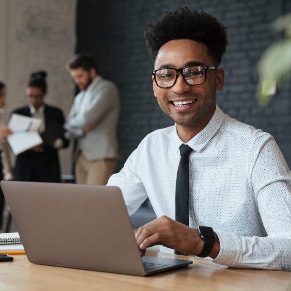 Photo of happy young african businessman sitting indoors using laptop computer. Looking camera.