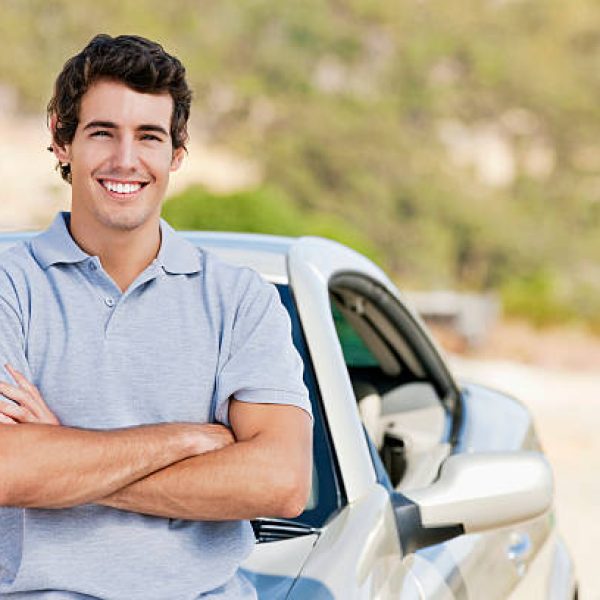 A young man smiles while standing in front of his car with his arms crossed.  Horizontal shot.